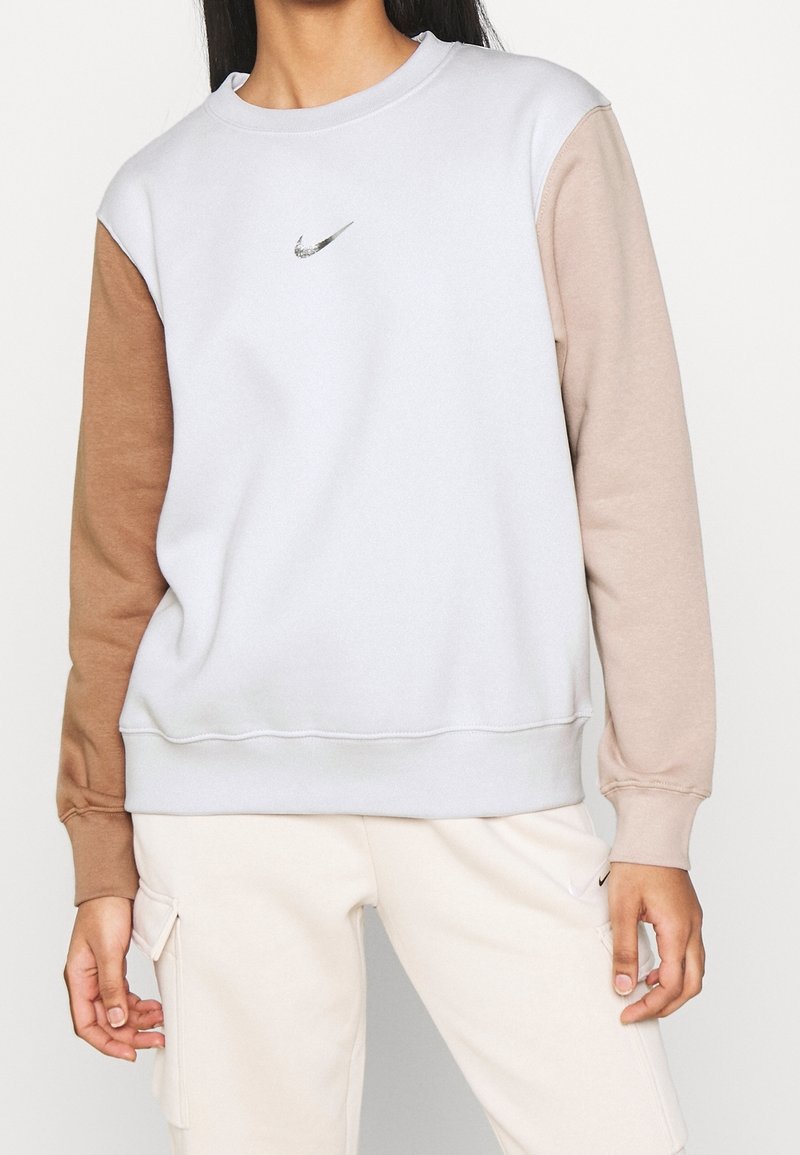 Femme portant un sweat-shirt blanc avec des manches beige et marron, un petit logo Nike sur la poitrine, et un pantalon cargo beige avec des poches.