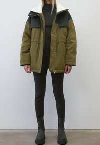 Olivegrüne Jacke mit schwarzen Schultern, weißem Fleece-Kragen, Frontreißverschluss und Kordelzug in der Taille. Schwarze Slim-Fit-Hosen und klobige schwarze Stiefel.