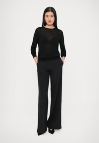 Μαύρο πλεκτό πουλόβερ με στρογγυλή λαιμόκοψη συνδυασμένο με μαύρο παντελόνι φαρδιάς γραμμής που διαθέτει ανάγλυφη λεπτομέρεια στο πλάι. Το μοντέλο στέκεται με τα χέρια στις τσέπες.