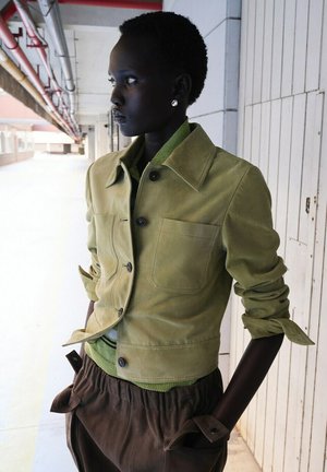 Szczupła osoba o krótkich włosach, ubrana w jasnozieloną kurtkę i brązowe spodnie, stojąca bokiem w korytarzu z kafelkami, z rękami w kieszeniach.