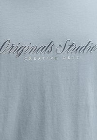 Υφασμα ανοιχτού γαλάζιου με το "Originals Studio" σε μεγάλη γραμματοσειρά και το "Creative Dept" σε μικρότερα κεφαλαία γράμματα από κάτω.