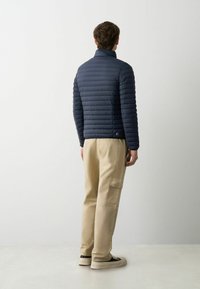 Doudoune marine avec un design matelassé horizontal, col montant et détail de logo en bas ; associée à un pantalon cargo beige et des baskets.