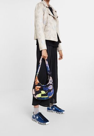 Personne portant une veste beige à motif floral, une jupe noire, des chaussettes argentées, des baskets bleues Mickey Mouse, tenant un sac à bandoulière noir avec un imprimé floral coloré.