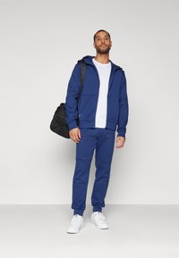 Marinblå sportig hoodie och byxor gjorda av mjukt tyg, med dragkedja fram och stora fickor, samt vita sneakers och en svart väska.