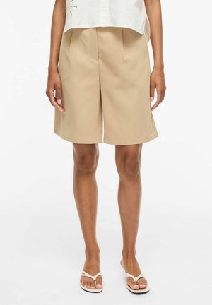 Kvinde iført beige knælange, skræddersyede shorts, hvid ærmeløs top og hvide flip-flops, stående på hvid baggrund.