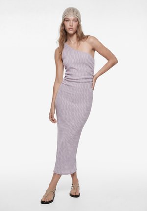 Kobieta w jasnofioletowej, prążkowanej sukience midi na jedno ramię, beżowej szydełkowanej czapce i beżowych sandałach, pozująca z jedną ręką na biodrze.