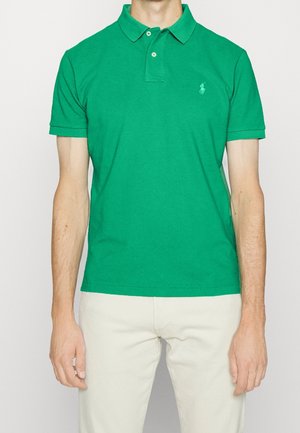Homme portant un polo vert vif à manches courtes avec deux boutons et un pantalon couleur crème, debout devant un fond uni.