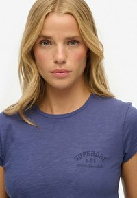 Camiseta azul con cuello redondo, mangas cortas y texto impreso "SUPERDAY NYC Athletic Essentials" en gris oscuro. Textura suave.