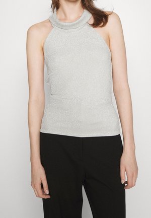 Kobieta w srebrnej bezrękawnikowej bluzce na szyję i czarnych spodniach, stojąca na jednolitym jasnym tle.