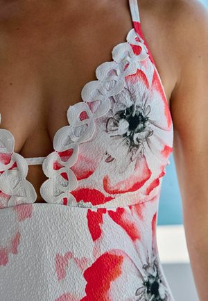 Zbliżenie na kobietę ubraną w teksturowaną białą sukienkę z czerwonymi i czarnymi kwiatowymi wzorami oraz białą koronką z falistym wykończeniem wzdłuż dekoltu.