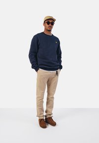 Sudadera azul marino con un pequeño logo naranja, pantalones beiges y zapatos marrones. Los accesorios incluyen una gorra color arena y gafas de sol redondas.