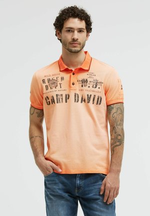 Man with curly hair wearing orange graphic polo shirt and blue jeans, standing with one hand in pocket against plain background.
