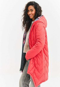 Prešitý kabát s kapucňou v jasnej koralovej farbe s kvetinovou vnútornou podšívkou. Má dlhé rukávy, šikmý lem a zapínanie na zips. Textúrovaná, teplá tkanina.