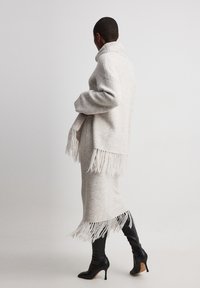 Weiches graues Strickoutfit mit hohem Kragen, verziert mit Fransenakzenten am Saum, kombiniert mit eleganten schwarzen High Heels.