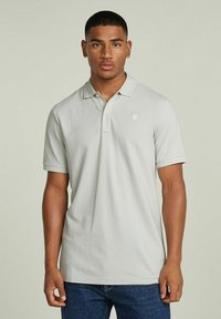 Camisa polo gris claro hecha de algodón suave, con un cierre de dos botones, mangas cortas y un pequeño logo en el pecho. Lleva puesta con jeans azules.