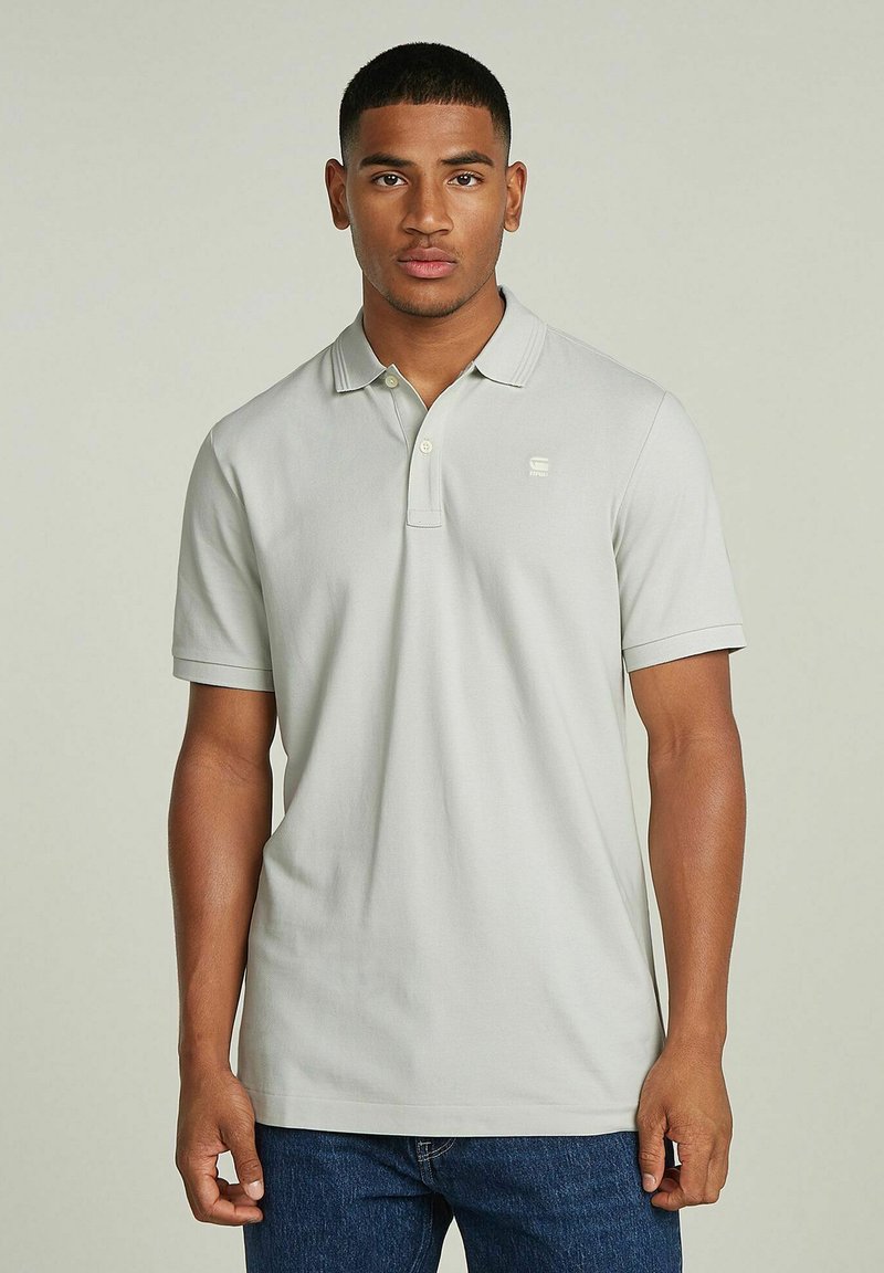 Camisa polo gris claro hecha de algodón suave, con un cierre de dos botones, mangas cortas y un pequeño logo en el pecho. Lleva puesta con jeans azules.