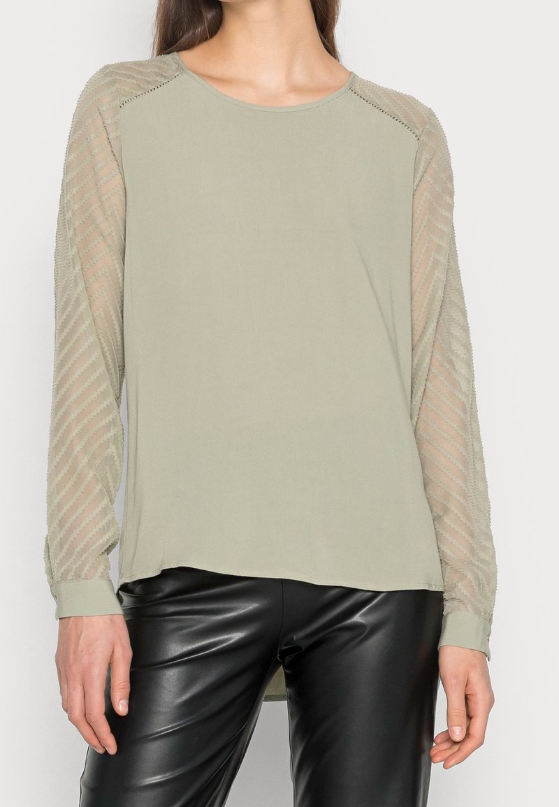 Blusa verde oliva chiaro con corpo uniforme e maniche lunghe trasparenti e stampate. Polsini a costine e scollatura rotonda. Texture liscia e vestibilità morbida.