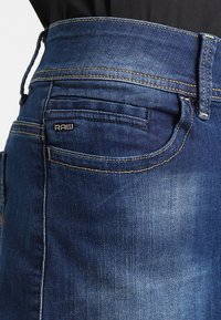 Jean en denim bleu foncé avec une texture délavée. Présente une poche arrière avec une étiquette noire "RAW" et des détails de surpiqûres en contraste.