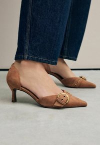 Scarpe basse in suede marrone con punta affusolata e una fascia decorativa con fibbia. Presentano un design senza retro e una suola texturizzata.