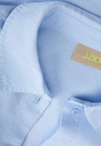 Lichtblauw overhemd met kraag, zichtbare knoop en beige neklabel met gele tekst op gladde stof.