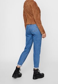 Veste en daim marron, jean en denim bleu avec des poignets retroussés et bottes à plateforme noires. Poches arrière visibles sur le jean. Design simple.