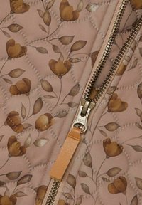 Tessuto trapuntato con un motivo floreale in tonalità terrose. Presenta una cerniera in metallo con un tirante in pelle color sabbia. Lo sfondo è di un rosa pallido.
