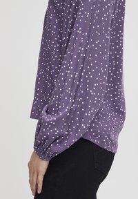 Lila långärmad topp med vita prickar, som har rynkade muddar och en avslappnad passform. Texturerad, elastisk tyg. Svarta byxor delvis synliga.