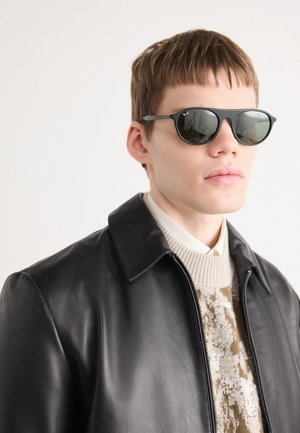 Mustad päikeseprillid ümmarguste läätsedega, hallist metallist raamiga, kannab mudel musta nahkjakki ja kudumit beežist pehmest materjalist, millel on mustrid.