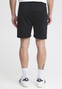 Pantalones cortos negros con cintura elástica, bolsillo con cremallera en la parte posterior y una textura suave, vistos desde la parte trasera. Calcetines blancos y zapatillas visibles en la parte inferior.