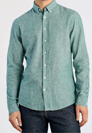 Homme portant une chemise bleu sarcelle à manches longues boutonnée avec un col et un jean bleu foncé, debout devant un fond uni.