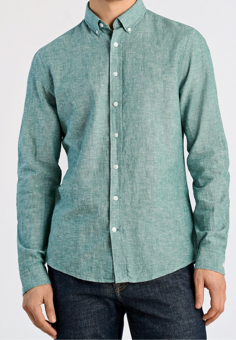 Homme portant une chemise bleu sarcelle à manches longues boutonnée avec un col et un jean bleu foncé, debout devant un fond uni.