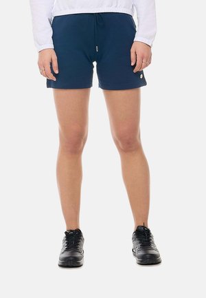 Persona che indossa pantaloncini blu navy, una giacca bianca e scarpe da ginnastica nere, in piedi con le mani rilassate ai lati.