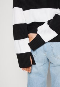 Pull à rayures noires et blanches en maille avec texture côtelée, fentes sur les côtés et manches longues. Associé à un jean en denim bleu clair.