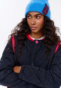 Žena s dlouhými kudrnatými vlasy, která má na sobě modrou pletenou čepici a prošívanou černou bundu s červenými detaily, hledí do strany se zkříženýma rukama.