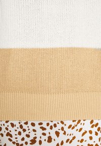Gros plan d’un pull en maille beige et blanc posé sur un tissu blanc avec des taches brunes irrégulières.