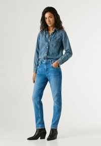 Vrouw staat en draagt een blauw denim shirt, ingestopt in medium blauwe jeans met zwarte enkellaarzen, hand in de zak, neutrale achtergrond.