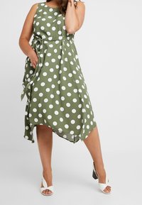 Grünes Kleid mit Tischlein in Punkten, runder Halslinie, tailliertem Bund, seitlichen Taschen und fließendem asymmetrischem Saum. Kombinieren Sie es mit weißen hochhackigen Sandalen.