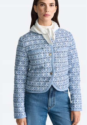Giacca in tweed blu con un motivo testurizzato, chiusura con bottoni e due tasche frontali, abbinata a una sciarpa bianca e jeans di denim.