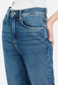 Jeans de mezclilla azul de talle alto con un acabado desgastado, cinco bolsillos, cierre de botón metálico y costuras naranjas a lo largo de las costuras.