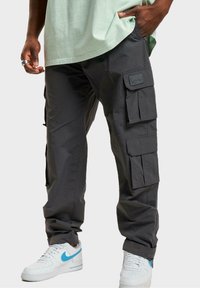 Cargo byxor i mörkgrått tyg, med flera sidofickor och en mjuk textur. Kombinerat med vita sneakers med ljusblå accenter.