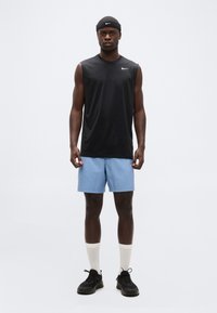 Αθλητής που φοράει μαύρο αμάνικο μπλουζάκι Nike, ανοιχτό μπλε σορτς, λευκές κάλτσες, μαύρα αθλητικά παπούτσια και μαύρη κορδέλα Nike στο κεφάλι, στέκεται όρθιος και κοιτάζει μπροστά.