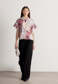 Žena kráčí v krátkém rukávu s abstraktním potiskem na blůze, širokých černých kalhotách, černých botách a drží malou světlou kabelku.