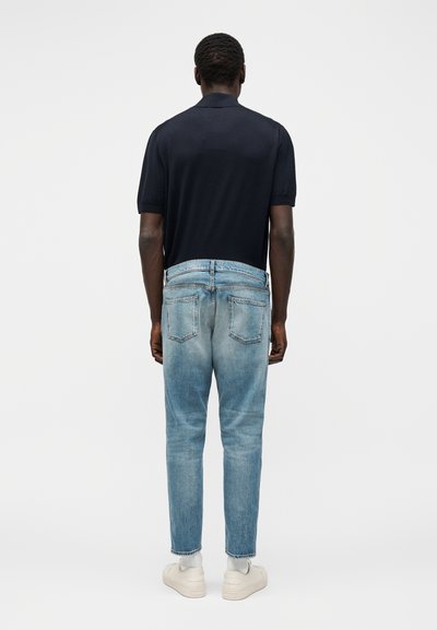 Homme portant une chemise noire à manches courtes, un jean bleu clair délavé, des chaussettes blanches et des baskets blanches, debout dos à la caméra.