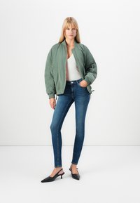 Green bomber jacket with ribbed collar, gray top, fitted blue jeans, and black slingback heels. Jacket features zip pockets and textured fabric.