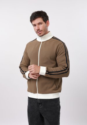 Moški si prilagaja rokave rjave jopice z zadrgo, belo ovratnikom in manšetami ter črnimi črtami na rokavih, oblečen v temne kavbojke, pred enostavnim ozadjem.