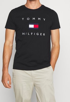 Mężczyzna ubrany w czarną koszulkę Tommy Hilfiger z logo w białym, czerwonym i granatowym kolorze, w połączeniu z jasnobeżowymi spodniami, stoi na jednolitym tle.