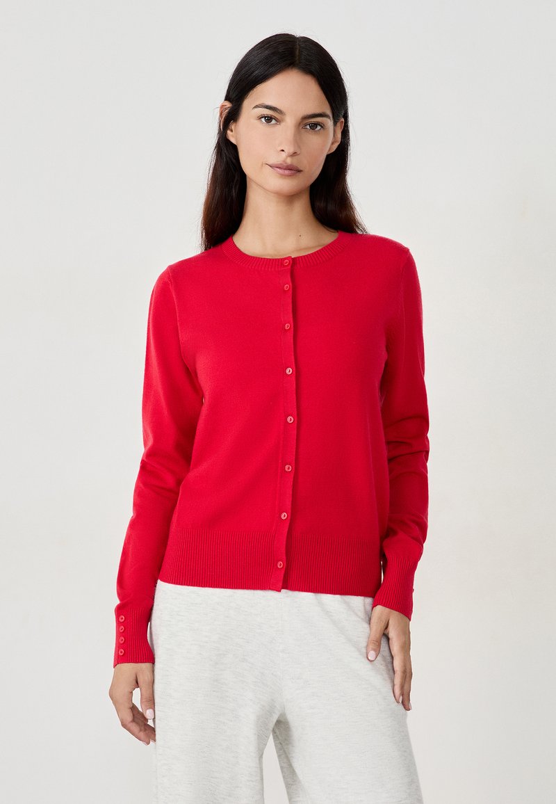 Cárdigan rojo de punto con cuello redondo, botones delanteros y puños y dobladillo acanalados. Combinado con pantalones gris claro. Textura suave.