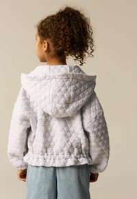 Hoodie gris matelassé avec une grande capuche, des poignets et un ourlet côtelés. Il présente un motif texturé et est associé à un jean bleu clair.