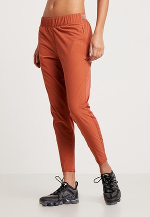 Frau trägt rostfarbene, schmal zulaufende Jogginghose und schwarze Sportschuhe, steht vor einem hellen Hintergrund.