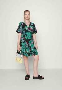 Vestido preto com padrões vibrantes de folhas verdes e rosas, mangas curtas em balão e saia com godê. Bolsa amarela e sandálias pretas tipo slide.